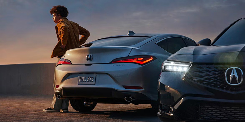 A person leaning against a silver 2025 Acura Integra with a black Acura Integra in the foreground at sunset.