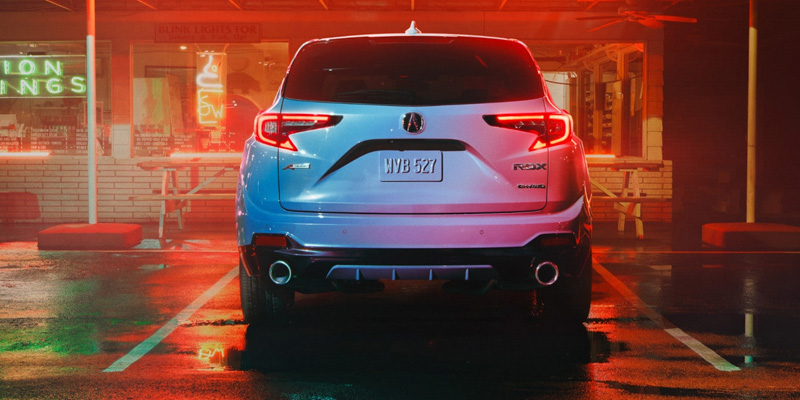 A white Acura RDX SUV is parked in a wet, neon-lit parking lot at night, viewed from the rear, with a restaurant in the background.